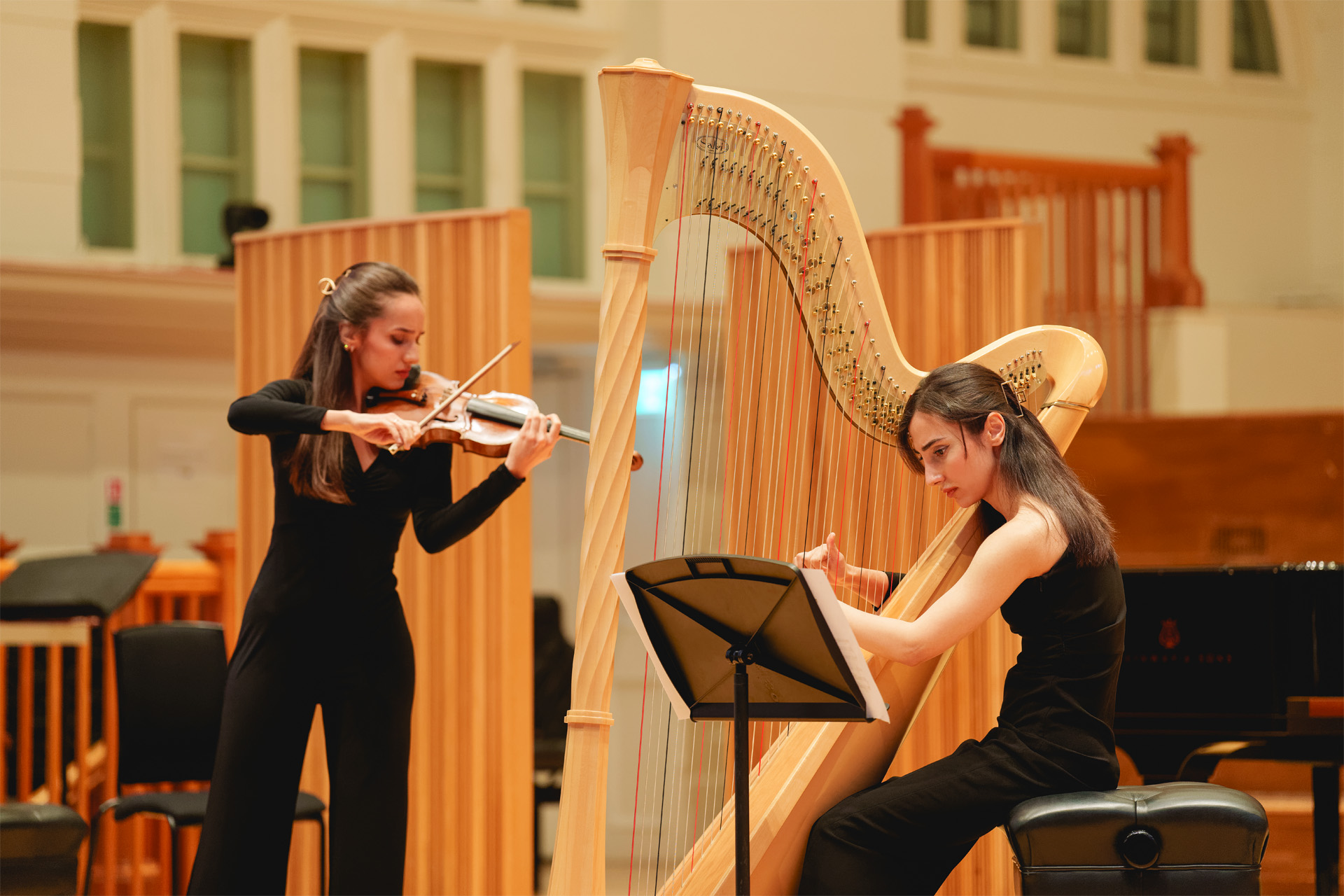 Two female students, wearing smart black attire, playing on a harp and violin on stage, with a wooden panelled background behind them.